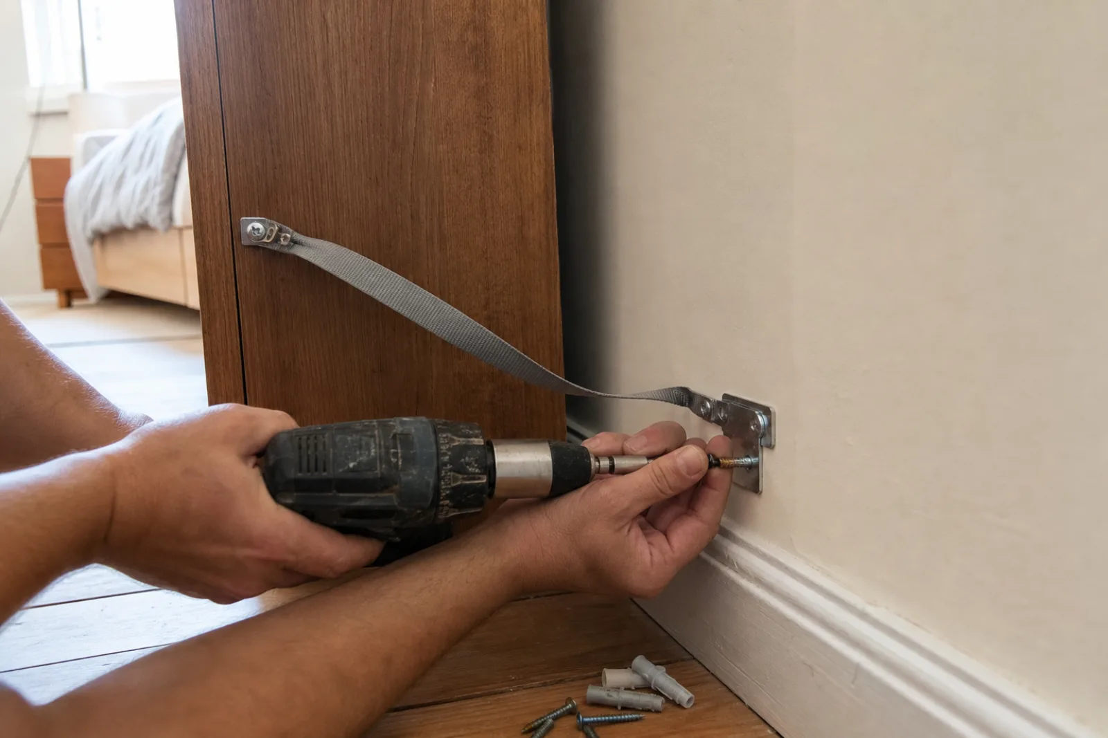Monteur schraubt eine Kippsicherung mit Spanngurt und Winkel von einem hohen Schrank an die Wand