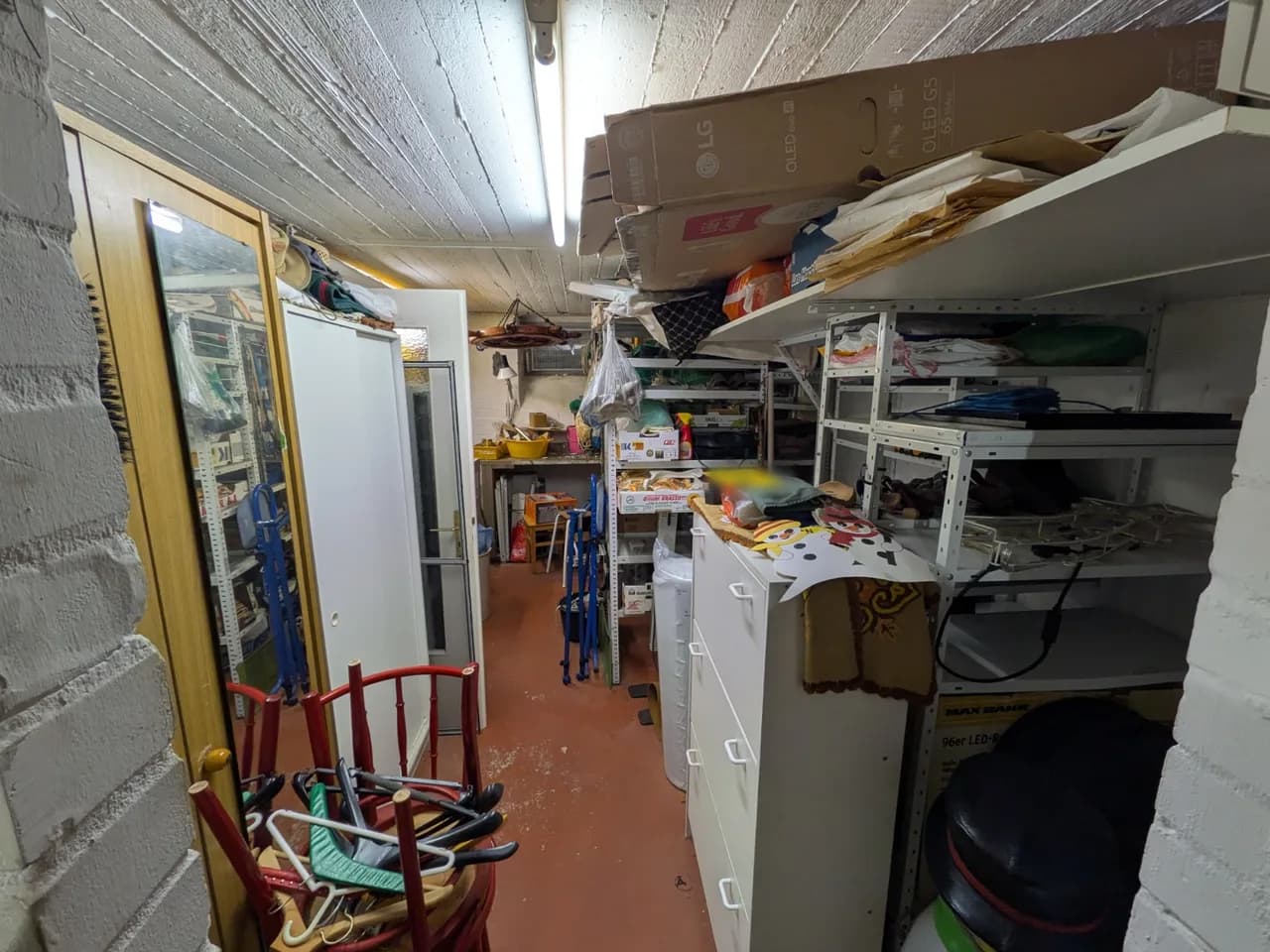 Blick durch den Keller in Fuhlsbüttel vor der Räumung: Kleiderschrank mit Spiegel, gestapelte rote Stühle, Kommode, Regale bis unter die Decke